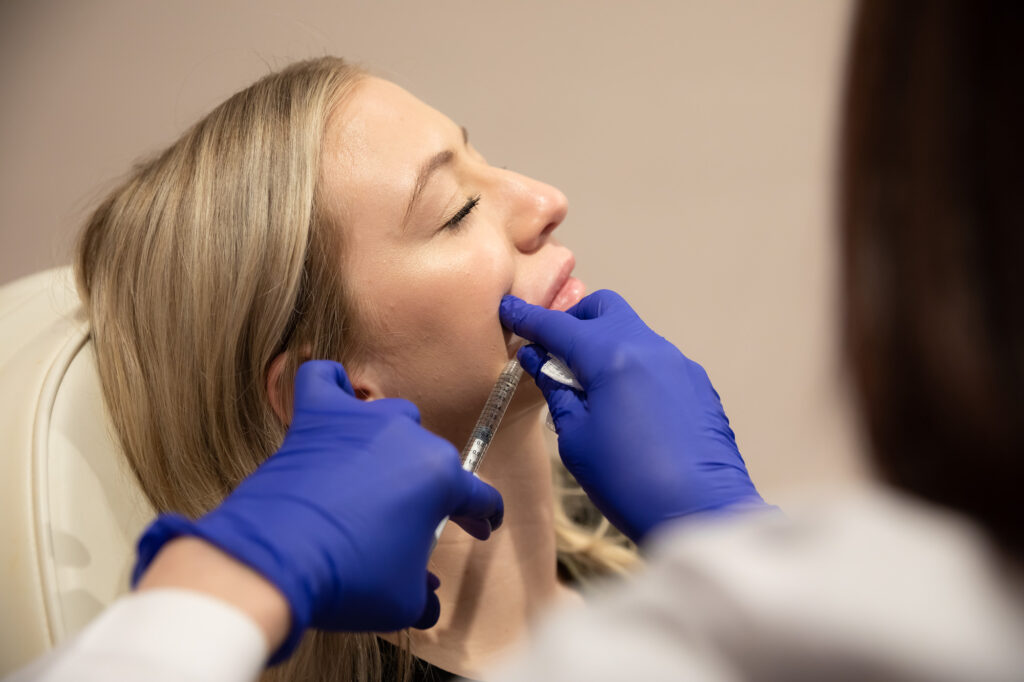 Woman lying down as gloved professional injects her lips with filler.
