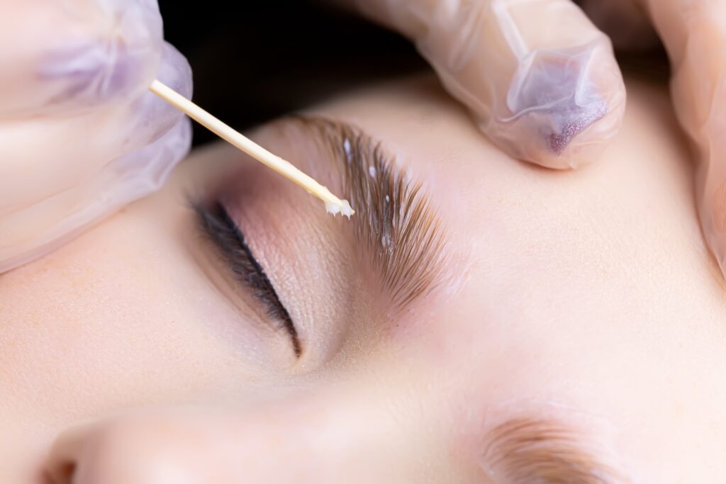 Practitioner wearing gloves applies brow tint with wooden stick.